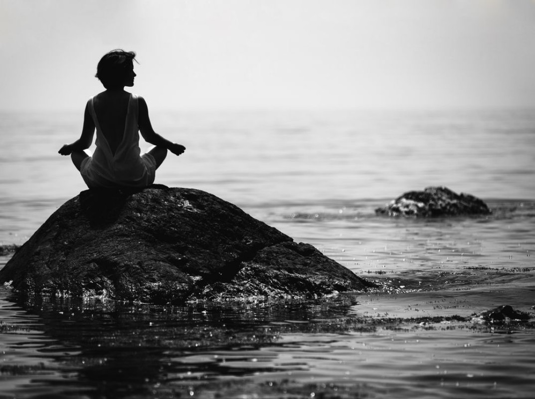 Picture of Young woman sits in a lotus position on a large stone in the sea and meditates her fingers are folded in Prithivi mudra